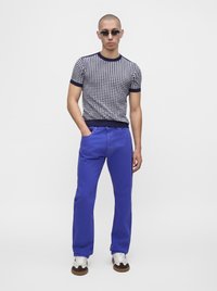 Homme portant des lunettes de soleil, une chemise à manches courtes à motifs géométriques, un pantalon bleu vif et des baskets blanches, debout avec une main dans la poche.