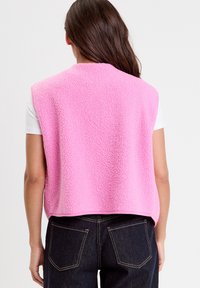 Pink fleece vest with a textured finish, sleeveless design, and a rounded hem, paired with a white shirt and dark denim jeans.