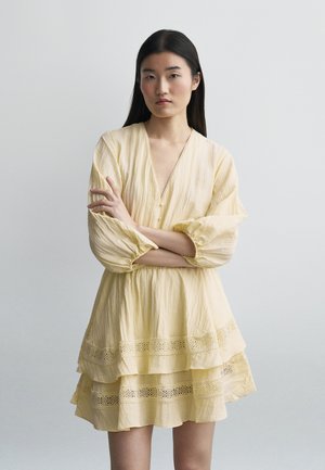 Fiatal nő hosszú sötét hajjal, réteges, világos sárga csipkerészletekkel díszített ruhában, karját keresztbe téve áll sima háttér előtt.