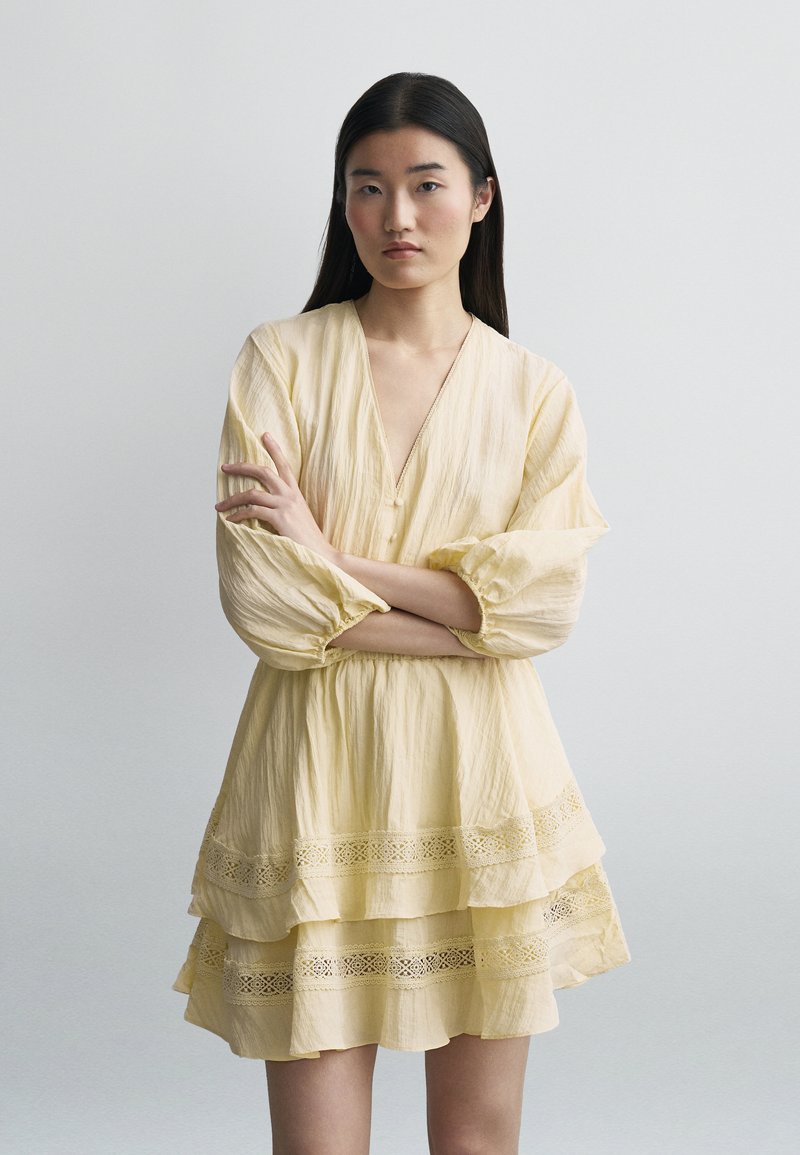 Νεαρή γυναίκα με μακριά σκούρα μαλλιά, ντυμένη με ένα ανοιχτό κίτρινο φόρεμα με στρώσεις και δαντελένες λεπτομέρειες, όρθια με σταυρωμένα τα χέρια μπροστά σε ένα απλό φόντο.