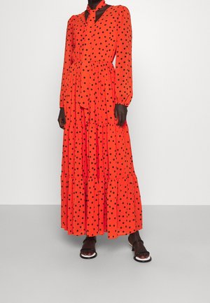 Kvinde iført en lang, lys orange kjole med sorte polkaprikker, en slipse detalje og lagdelt nederdel, kombineret med sort/hvide sandaler.