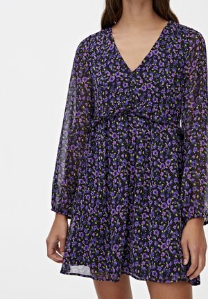 Kvinde iført en gennemsigtig sort kjole med lange ærmer og lilla blomsterprint, med V-udskæring og knapper ned foran.