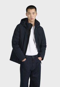 Námořnická puffer bunda s kapucí, předním zipem a logem. Model ji kombinuje s bílým tričkem a tmavě modrými džíny.