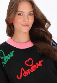 Jonge vrouw met lang bruin haar, draagt rode oordopjes en een zwarte trui met een roze kraag en kleurrijke "Amour" tekstontwerpen.