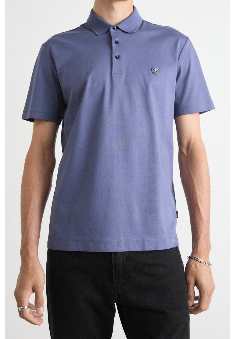 Polo tričko s krátkým rukávem v jemné fialové barvě, vybavené klopovým límečkem na tři knoflíky a vyšitým logem na hrudi. V kombinaci s černými kalhotami.