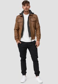 Veste bomber en cuir marron avec fermeture éclair, doublure à capuche grise et quatre poches à l'avant, associée à un pantalon noir fuselé et des baskets blanches.