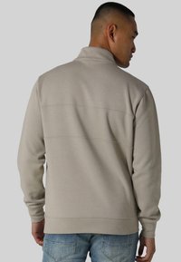 Lys beige sweatshirt med høy krage, med teksturert stoff og sømdetaljer. Kombinert med dongeribukser, sett fra baksiden.