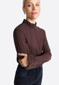 Rich brown long-sleeve top with a ruffled high neck and cuffs, featuring a soft texture and fitted design. Paired with dark jeans.