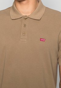 Beige polotröja i strukturväv, med krage och placket med tre knappar, samt en röd Levi's-logotyp på bröstet.