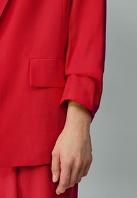 Rode blazer van soepel materiaal, met een voorste patchzak en opgerolde manchetten. Close-up van de mouw en een hand.