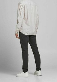 Muž v světle béžové košili s dlouhým rukávem, černých slim-fit kalhotách a bílých teniskách stojící zády k fotoaparátu na šedém pozadí.