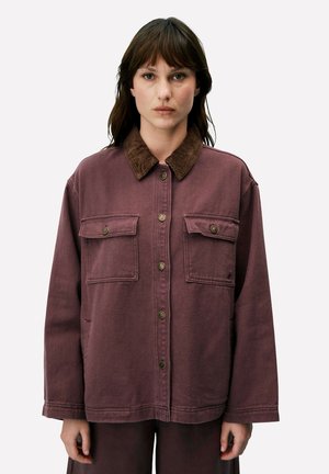 Vrouw draagt een loszittende, paarse jas met knopen en een bruine ribfluwelen kraag, staand tegen een effen witte achtergrond.