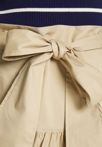 Beiger Stoffgürtel mit einer Schleifendetaillierung, über einem gerafften Rockteil. Dunkelblause Oberteil mit weißen Streifen ist teilweise sichtbar.