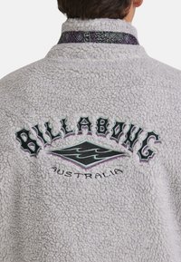 Billabong fleecejakke, lys grå, struktureret stof, broderet logo i sort og lilla. Dekorativ mønstret krave.
