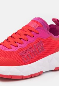 Baskets rouge vif et rose avec une tige en maille texturée, des lacets rouges, une semelle blanche à motif géométrique et le logo "Versace Jeans Couture" sur le côté.