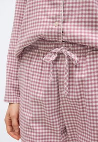 Γυναίκα που φοράει ροζ και λευκή πιτζάμα με καρό σχέδιο, σφιχτή μέση με κορδόνι και κουμπωτό πάνω μέρος, με το χέρι χαλαρό στο πλάι.