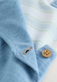 Col de chemise bleu clair avec bouton marron et boutonnière brodée, posé sur un tissu blanc à rayures vert pâle et bleues.