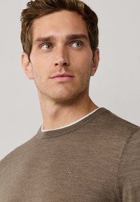 Lichtgewicht bruine trui met een geribbelde halslijn en subtiele textuur. Bevat een klein geborduurd logo op de borst.