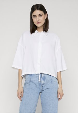 Kvinde med glat mørkt hår iført løs hvid skjorte med knapper og lyseblå højtaljede jeans, stående mod en ensfarvet baggrund.