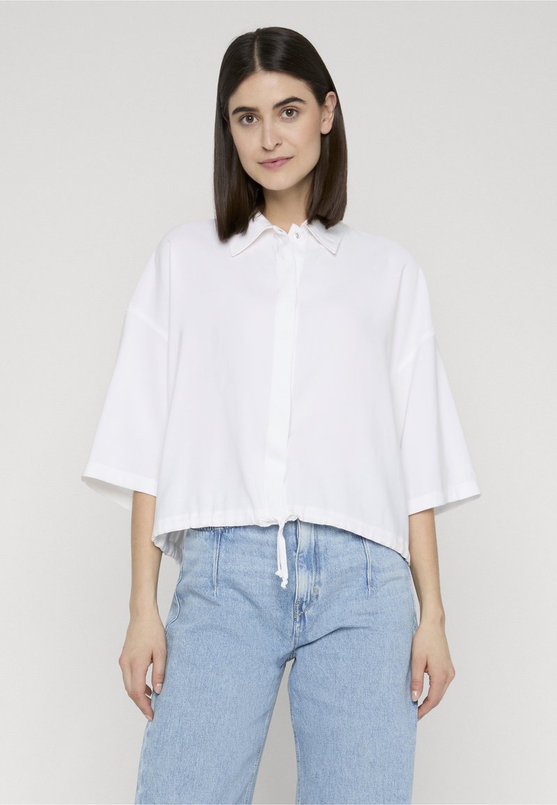 Femme aux cheveux raides et foncés, portant une chemise blanche ample à boutons et un jean taille haute bleu clair, se tenant devant un fond uni.