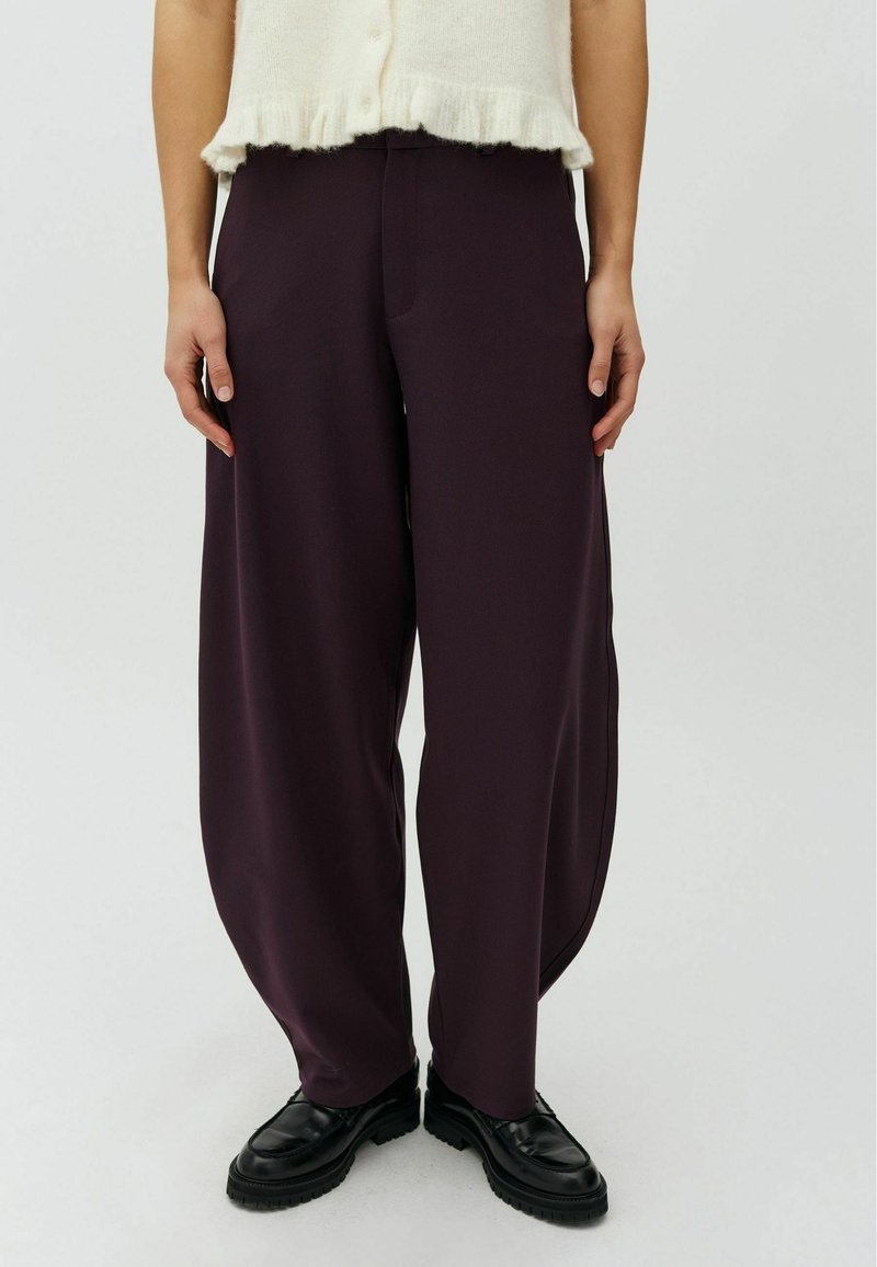 Personne portant un pantalon ample violet foncé, des chaussures noires et un pull court crème, debout devant un fond uni.