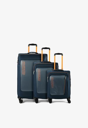 Tre valigie soft-shell blu navy con manici telescopici e ruote arancioni, etichettate "American Tourister", mostrate in dimensioni piccola, media e grande.