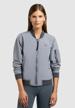 Kobieta w szarej bomberce z suwakiem i detalem kieszeni na rękawie, stojąca na jednolitym jasnym tle.