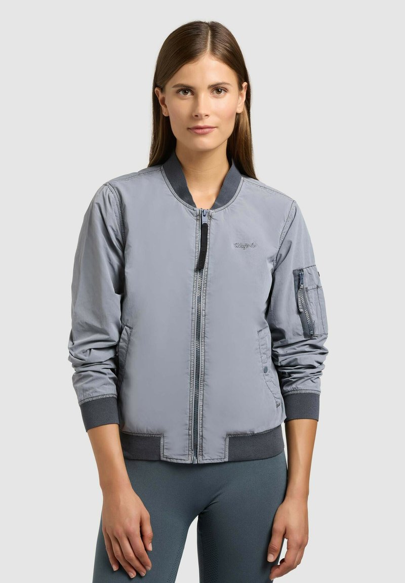 Femme portant une veste bomber grise avec fermeture éclair et détail de poche sur la manche, debout devant un fond clair uni.