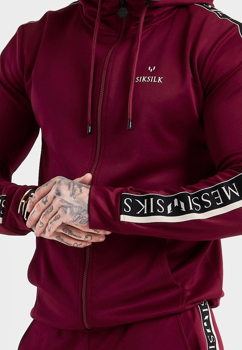 Burgunderfarbener Zip-Hoodie mit hohem Kragen, ausgestattet mit schwarzem und cremefarbenem Logoband an den Ärmeln, aus einem glatten, dehnbaren Material gefertigt.