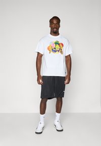 Hvit kortermet t-skjorte med fargerik grafikk; kombinert med svarte shorts og hvite sneakers; ensfarget bakgrunn.