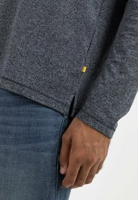 Grå langærmet skjorte med en tekstureret overflade, der har en sideslids og et lille gult accentmærke, båret med blå denimjeans.