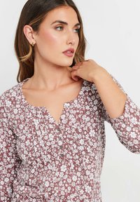 Bluse mit floralem Muster in Malve und weißen Blumen, runde Ausschnittform und drei Knöpfe. Mit Falten und langen Ärmeln. Strukturierter Stoff.