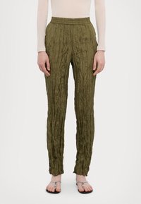 Persoon in olijfgroene gekreukte broek, doorzichtige beige lange mouw top en zilveren teensandalen, staand tegen een effen achtergrond.