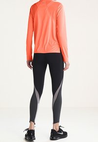 Helles orangefarbenes, langärmeliges Sportoberteil mit rundem Ausschnitt, kombiniert mit schwarzen Leggings mit grauen Seitenpaneelen und einer strukturierten Oberfläche.