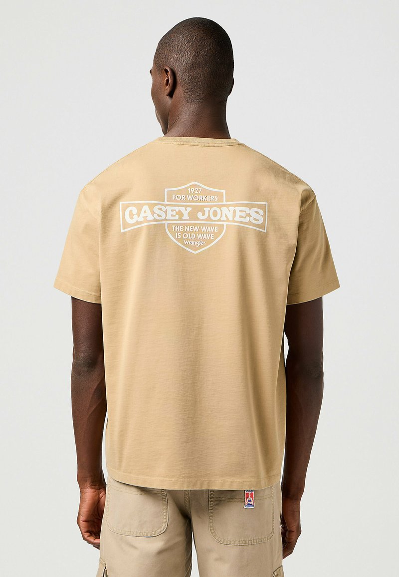 Camiseta de manga corta color marrón con un corte relajado, que presenta texto impreso en blanco en la parte posterior, incluyendo "CASEY JONES" y una etiqueta con el logo en el dobladillo.