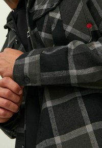 Camicia in flanella a quadretti verdi e neri con superficie testurizzata, dotata di un polsino con bottone e accenti rossi. Visione ravvicinata del tessuto e del design.
