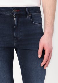 Tummansiniset farkut, joissa on slim fit -leikkaus, vetoketjukiinnitys, punaiset korost stitchingit ja perinteinen viisi taskua -suunnittelu.