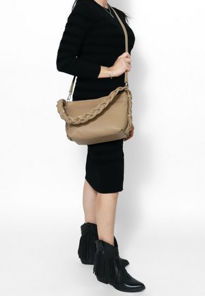 Borsa a spalla in pelle beige con tracolla intrecciata, texture morbida, forma rettangolare e fondo piatto, tenuta da una persona che indossa un vestito nero aderente.