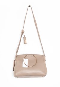 Béžová kožená kabelka přes rameno s nastavitelným popruhem, velkým zlatým kulatým úchopem na přední straně a připojenou béžovou visačkou se zlatým písmem.