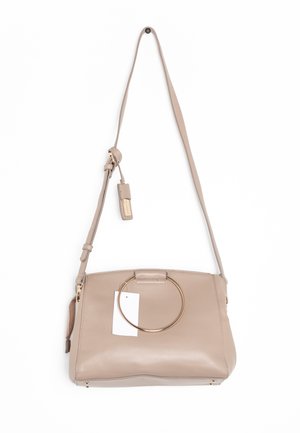 Béžová kožená kabelka přes rameno s nastavitelným popruhem, velkým zlatým kulatým úchopem na přední straně a připojenou béžovou visačkou se zlatým písmem.