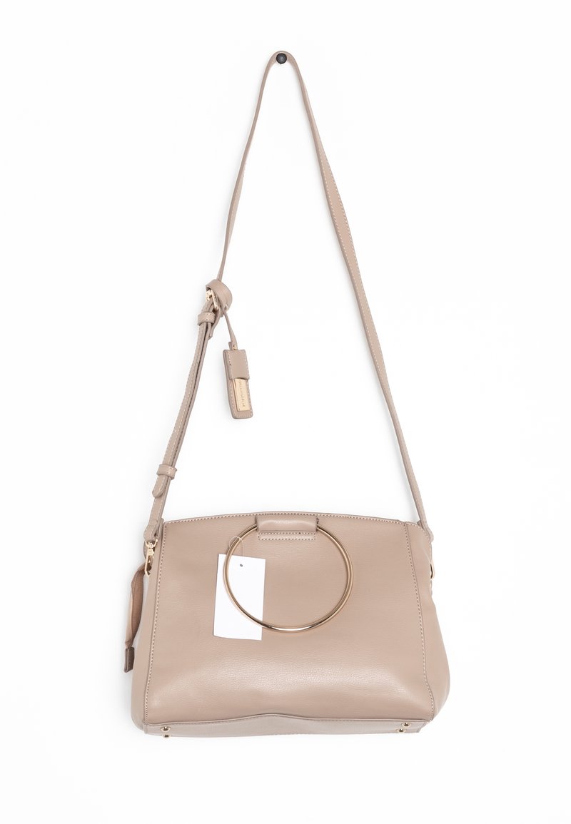 Béžová kožená kabelka přes rameno s nastavitelným popruhem, velkým zlatým kulatým úchopem na přední straně a připojenou béžovou visačkou se zlatým písmem.