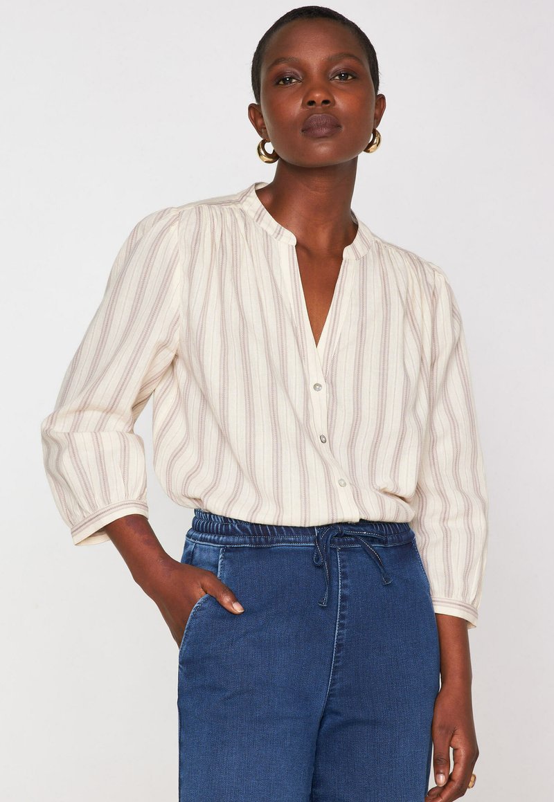 Donna che indossa una blusa a righe crema con maniche a trequarti e pantaloni blu a vita alta con cordoncino, mano nella tasca.