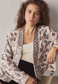 Sequin-embellished jacket with a sheer fabric; features an open front, soft beige color, and intricate silver and brown patterns.