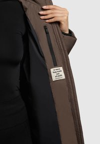 Braune isolierte Jacke mit schwarzem Innenfutter, ausgestattet mit einer Reißverschlusstasche und einem Stoffetikett, auf dem steht: "Eine warme Umarmung aus Stoff."