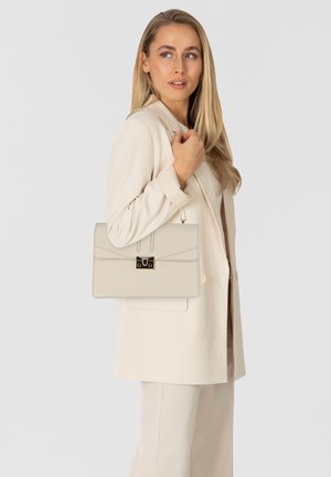 Sac à main en cuir beige avec un design à rabat, matériel doré et une sangle en chaîne, tenu par une personne portant une tenue beige assortie.