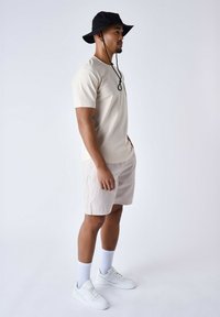 Camiseta beige con un logo, pantalones cortos beige claro, zapatillas blancas y un sombrero de cubo negro con una correa para el mentón. Presentado sobre un fondo sencillo.