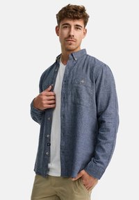 Camisa de hombre de botones en tejido texturizado azul marino, con un bolsillo en el pecho, cuello clásico y botones marrones, superpuesta sobre una camiseta blanca.