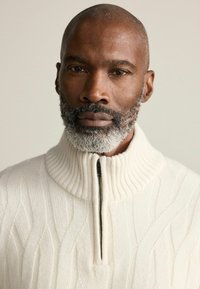 White, ribbed knit sweater with a high collar and half-zip, showing a textured pattern. Model has a close-shaved head and gray beard.