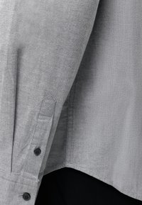 Gros plan sur un poignet de chemise grise à manches longues avec des boutons noirs et des coutures visibles sur un pantalon noir.