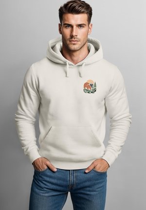 Young man wearing a cream hoodie with a small nature design on the chest, standing with hands in pockets against a plain gray background.